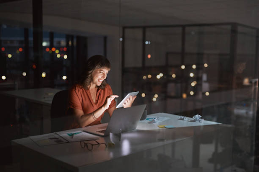 A woman works late in an office, using a tablet and laptop, symbolizing decisions in app development tech stack.